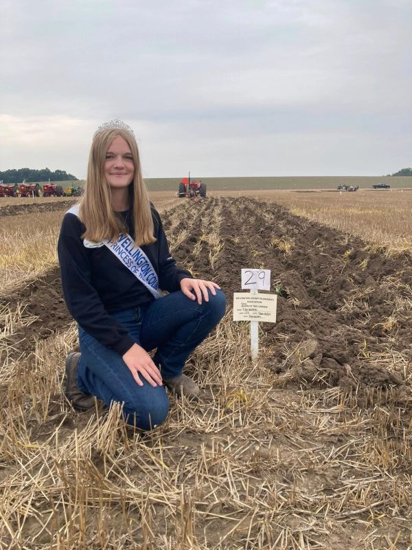 Young woman with a crown and sash kneeling in a recently plowed field.