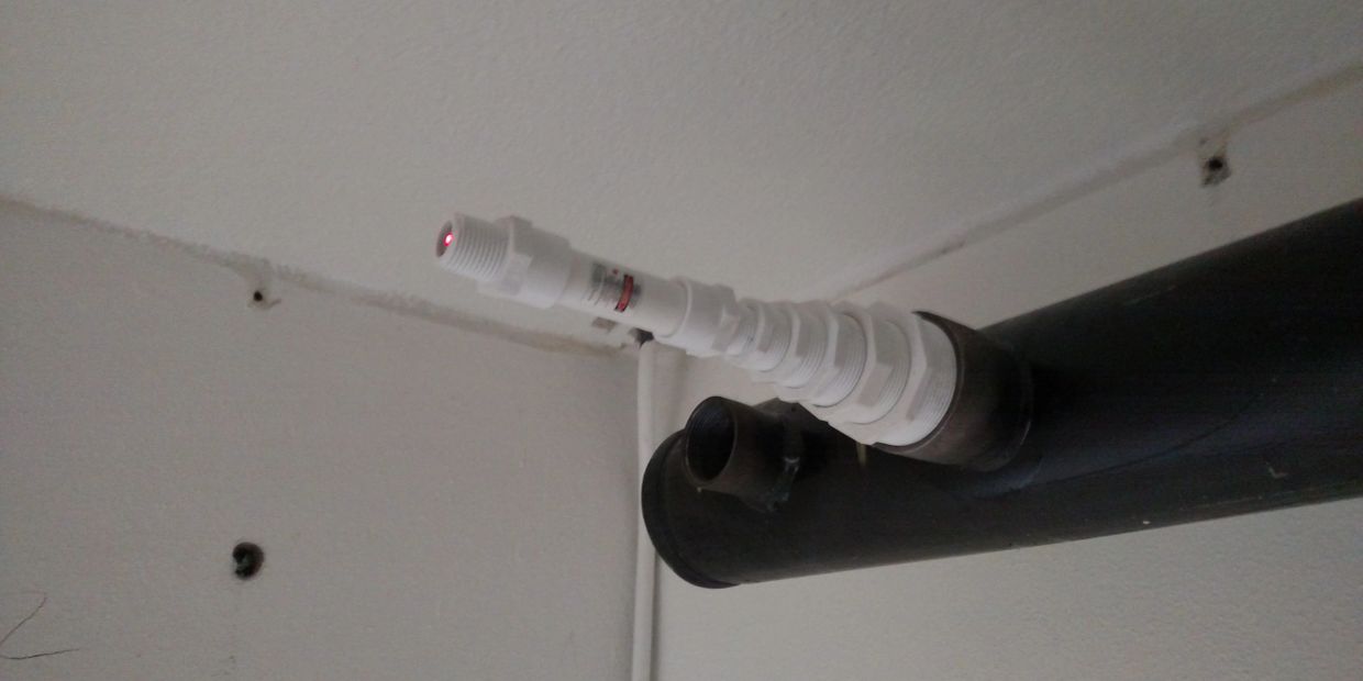 White laser device emitting red light attached to a black pipe on a wall.