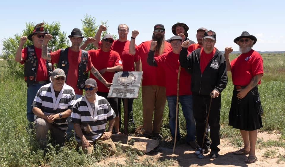 Group of men raising fists around a commemorative plaque in a grassy field.