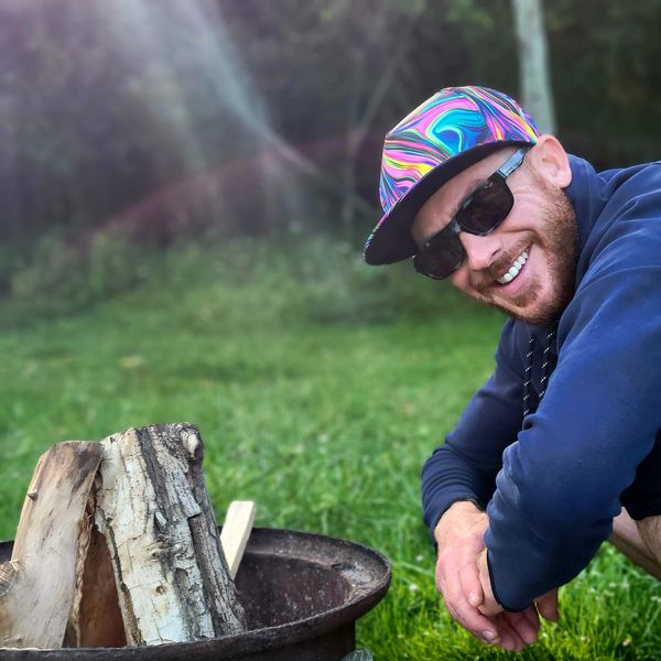Man smiling outdoors wearing sunglasses and a colorful cap near a fire pit.