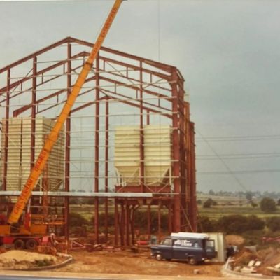 Steel structure under construction with cranes and vehicles on a rural site.
