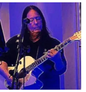 Man with long hair playing an acoustic guitar and singing into a microphone on stage.