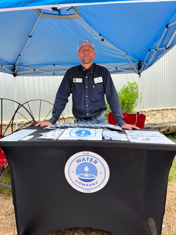Man stands behind a promotional table for The Water Company under a blue canopy.