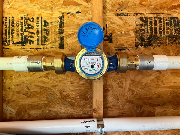 Newly installed water meter with labeled pipes on wooden wall.