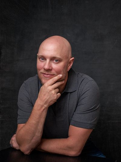 Smiling bald man in a gray polo shirt posing thoughtfully against a dark background.