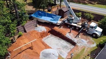 A crane lifts a blue pool into a backyard excavation site.