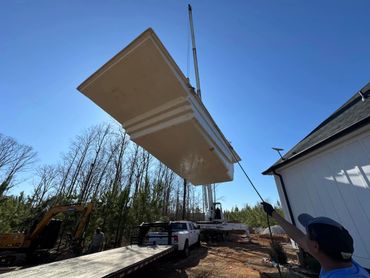 A crane lifts a large pool shell while a worker guides it with a pole.