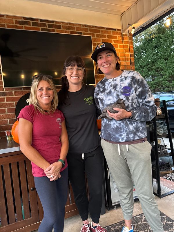 Three women smiling together on a patio, one holding a small bird.