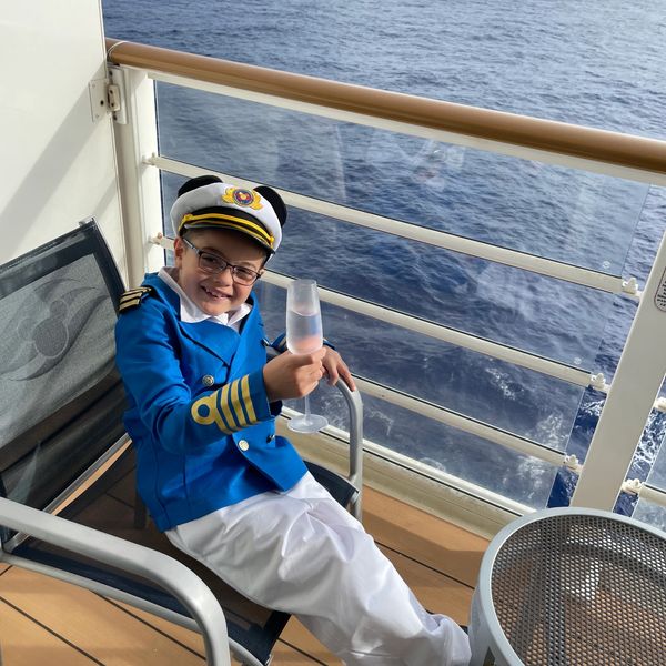 A smiling boy in a captain's uniform relaxing on a ship balcony with a drink.