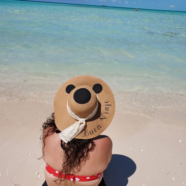 Woman in a red polka dot bikini and sunhat sitting on a beach by clear blue water.