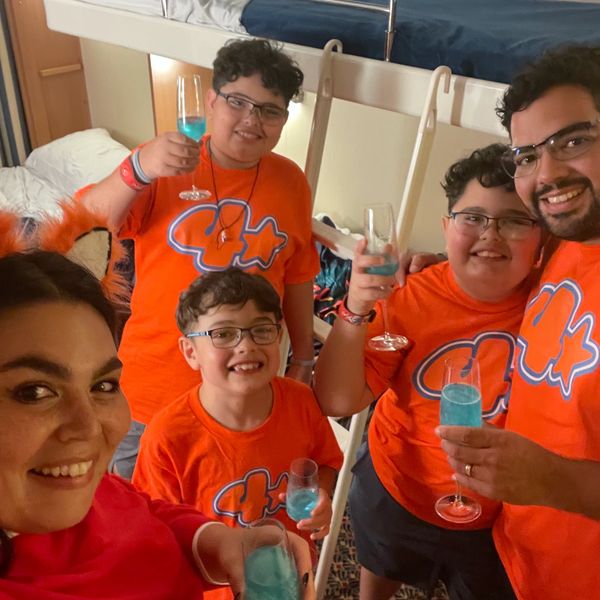 Family of five wearing matching orange shirts, smiling and toasting with blue drinks in a cozy room with bunk beds.