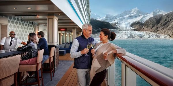 Couple toasting with drinks on a cruise ship with snowy mountains in the background.