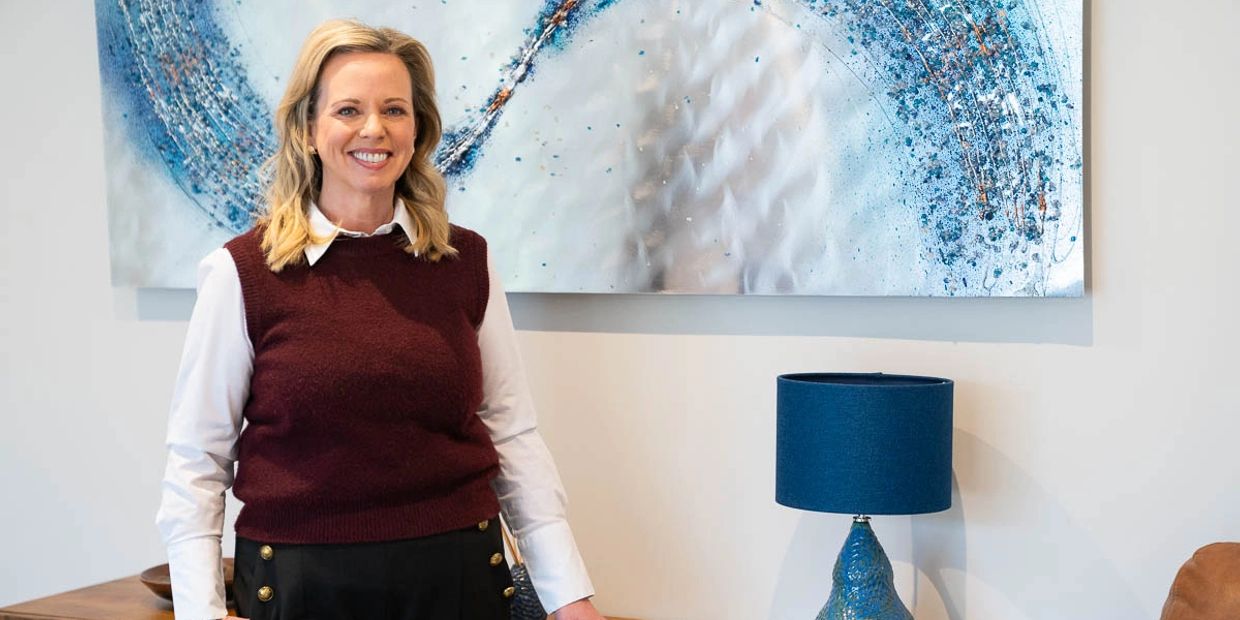 Smiling woman in a burgundy sweater vest stands by a blue lamp and abstract wall art.