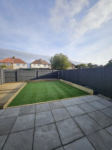 A backyard with a tiled patio, artificial grass, and dark wooden fences.