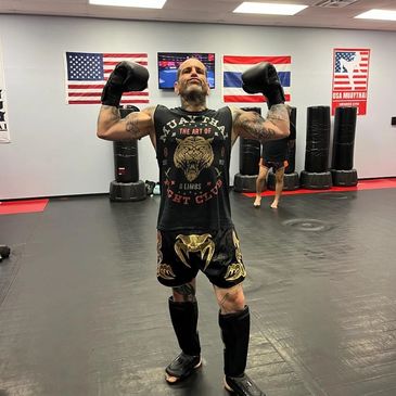 Man flexing muscles in a gym with boxing gloves and Muay Thai shorts.