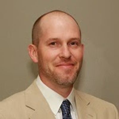 Man in beige suit and tie smiling against a plain background.