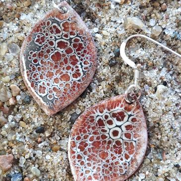 Close-up of handcrafted vitreous enamel earrings with intricate rust and white patterns on sandy background.