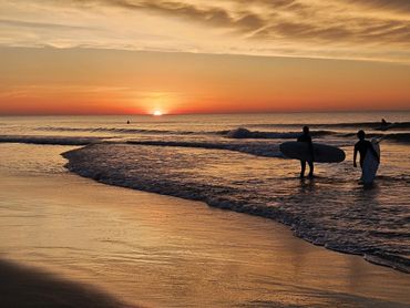 Surfers walking into the ocean at sunset with vibrant orange skies.