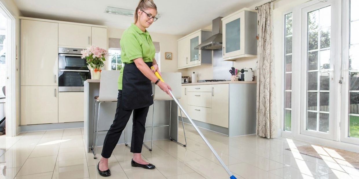 Woman mopping floor in modern kitchen with large windows and light tiles.