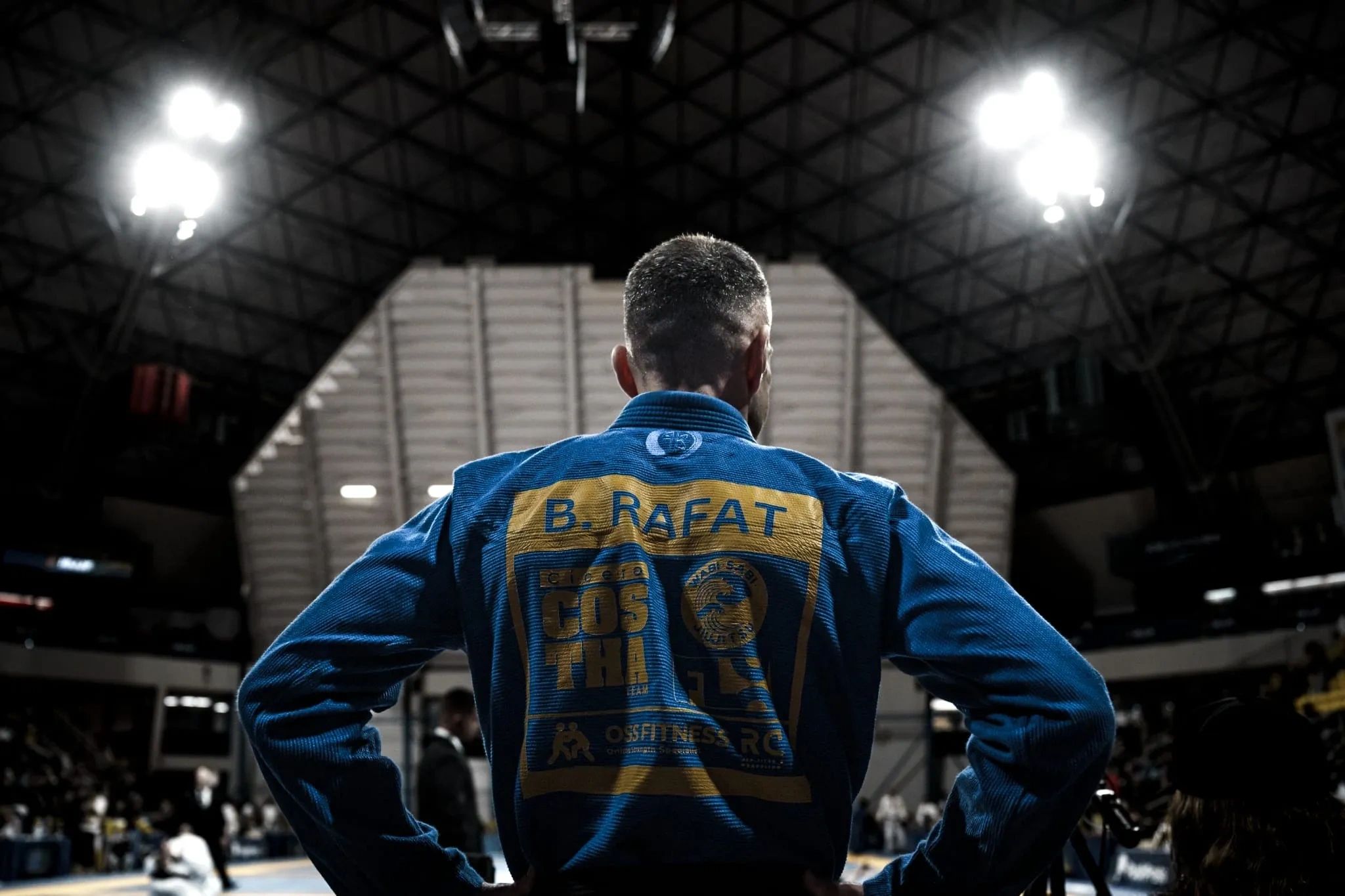 Athlete in a blue gi stands with hands on hips in an indoor arena.