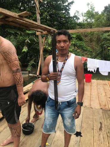 Man holding a traditional spear in a rustic outdoor setting with tribal tattoos visible on another person.