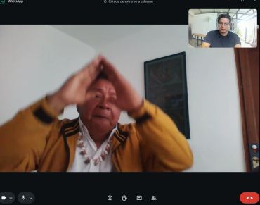 Two men video calling, one wearing a yellow jacket making a hand gesture.