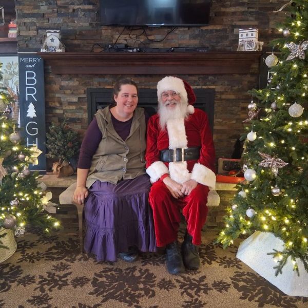 A woman sitting next to Santa Claus between two decorated Christmas trees.