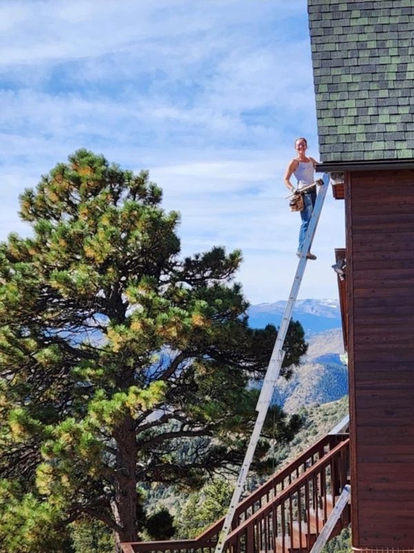 A person on a tall ladder working on a house roof beside a large tree.