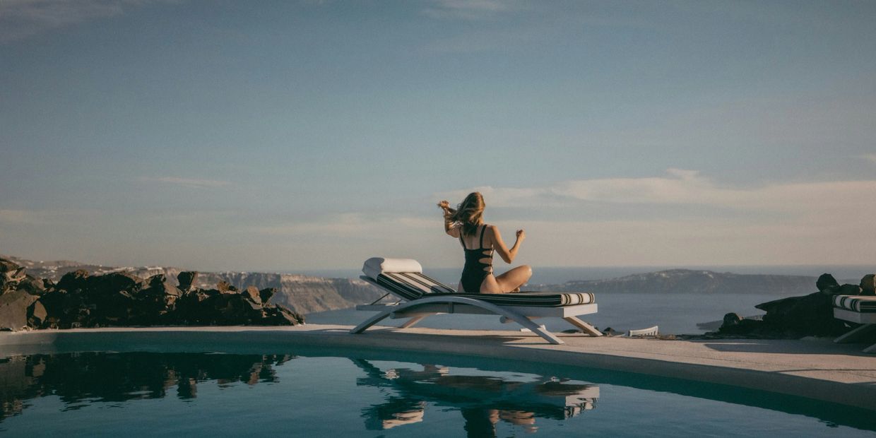 Woman in swimsuit sitting on a poolside lounge chair overlooking the sea.