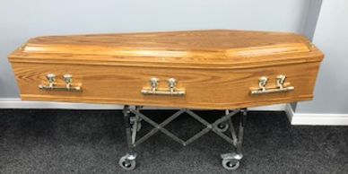 An oak veneered, raised lid coffin with plain sides.  Fitted with Oscar handles and finished in a me