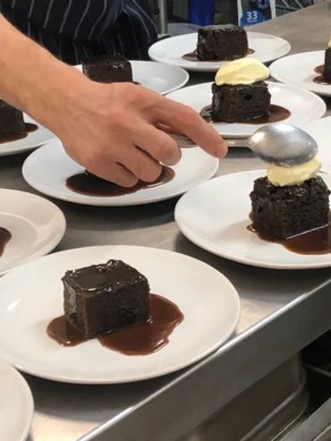 Plating chocolate cake squares with cream on white plates in a kitchen.