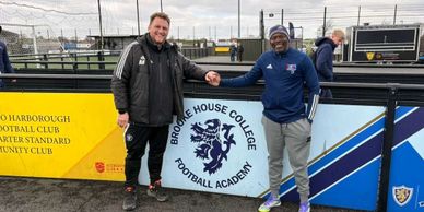 Two men shaking hands in front of a football academy sign on a sports field.