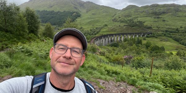 Man takes a selfie with a train viaduct in the background.