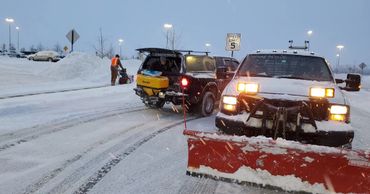 Two trucks and a person clearing snow in a snowy parking lot at dusk.