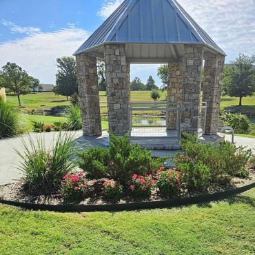 Stone gazebo with metal roof in a sunny park surrounded by greenery and flowers.