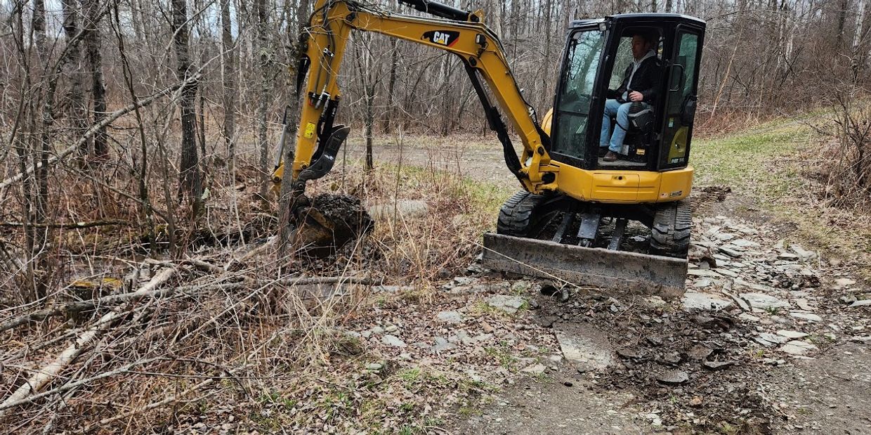 A person operates a small yellow excavator on a rocky, wooded path.