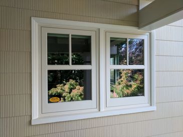 White-framed double windows reflecting greenery outside on beige siding.