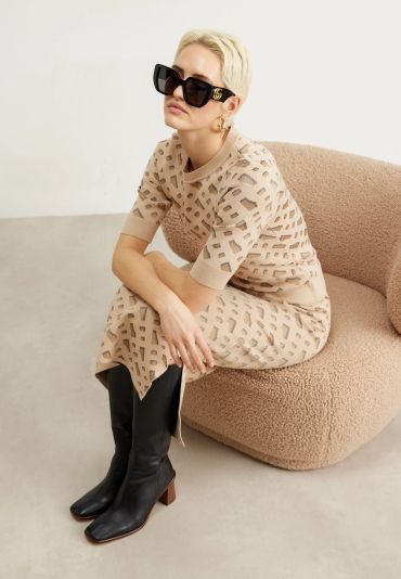 Stylish woman in beige patterned dress and large sunglasses sitting on a textured chair.
