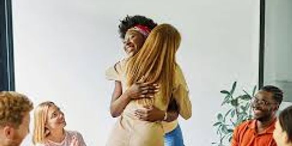 Two women warmly hugging in a group setting with others smiling.
