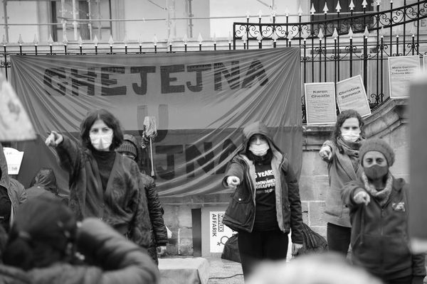 Women wearing masks point forward during a protest with a red banner behind them.