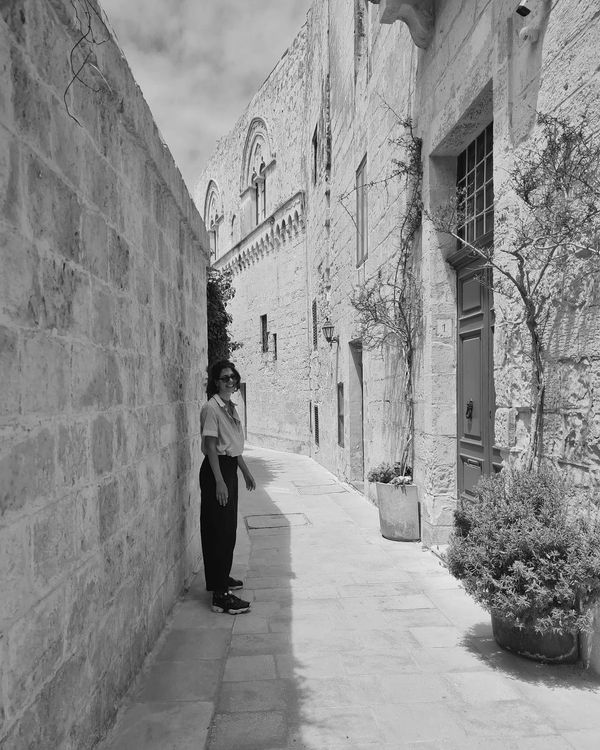 Woman standing in a sunny narrow stone alley with blue door and potted plants.