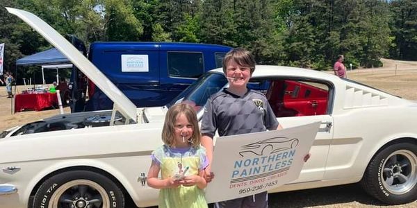 Two kids posing with a classic white car at an outdoor event.