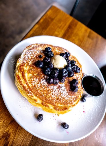 Stack of pancakes topped with blueberries, butter, and powdered sugar, served with syrup.