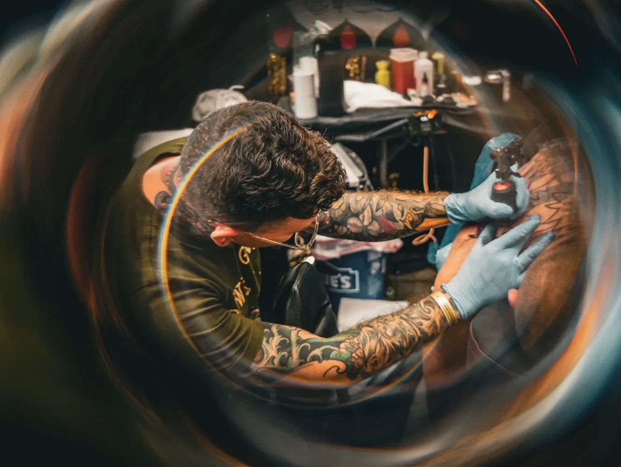 Tattoo artist with tattooed arms working on a client's arm in a studio.