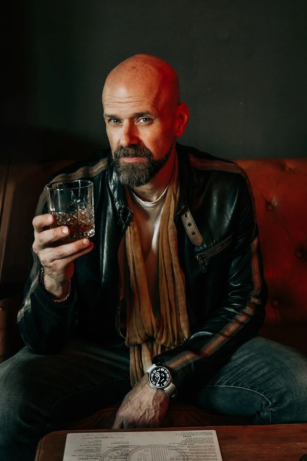 Bald man with beard holding a glass of whiskey, sitting on a leather couch.