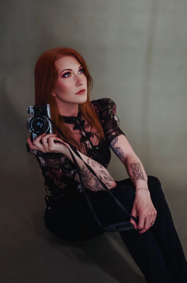 Red-haired woman with tattoos holding a camera, looking thoughtfully upwards.