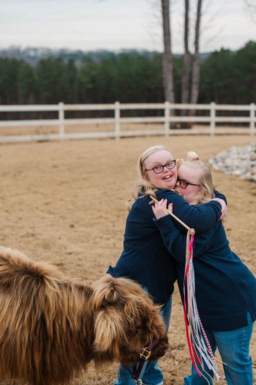 Two women hugging outdoors near a small brown Highland cow.