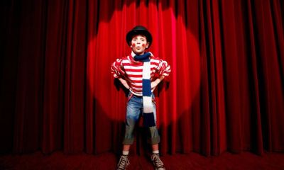 A child dressed as a mime stands on stage in front of red curtains.