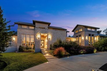 Contemporary stone home with an illuminated entry and landscaped driveway at twilight