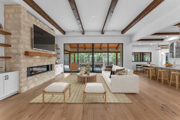 Open-plan living room with exposed wood beams, a stone fireplace and patio views